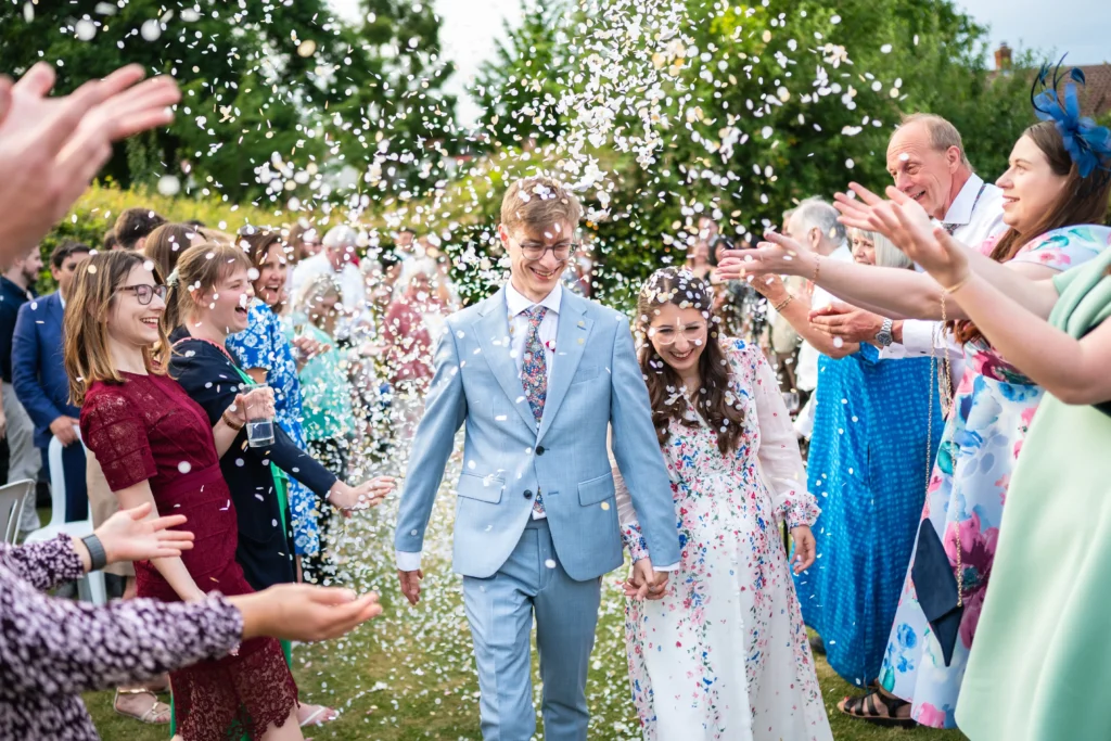 Bride in a floral wedding dress and groom in a light blue suit walking through a confetti shower with cheering wedding guests, Nottingham wedding photographer