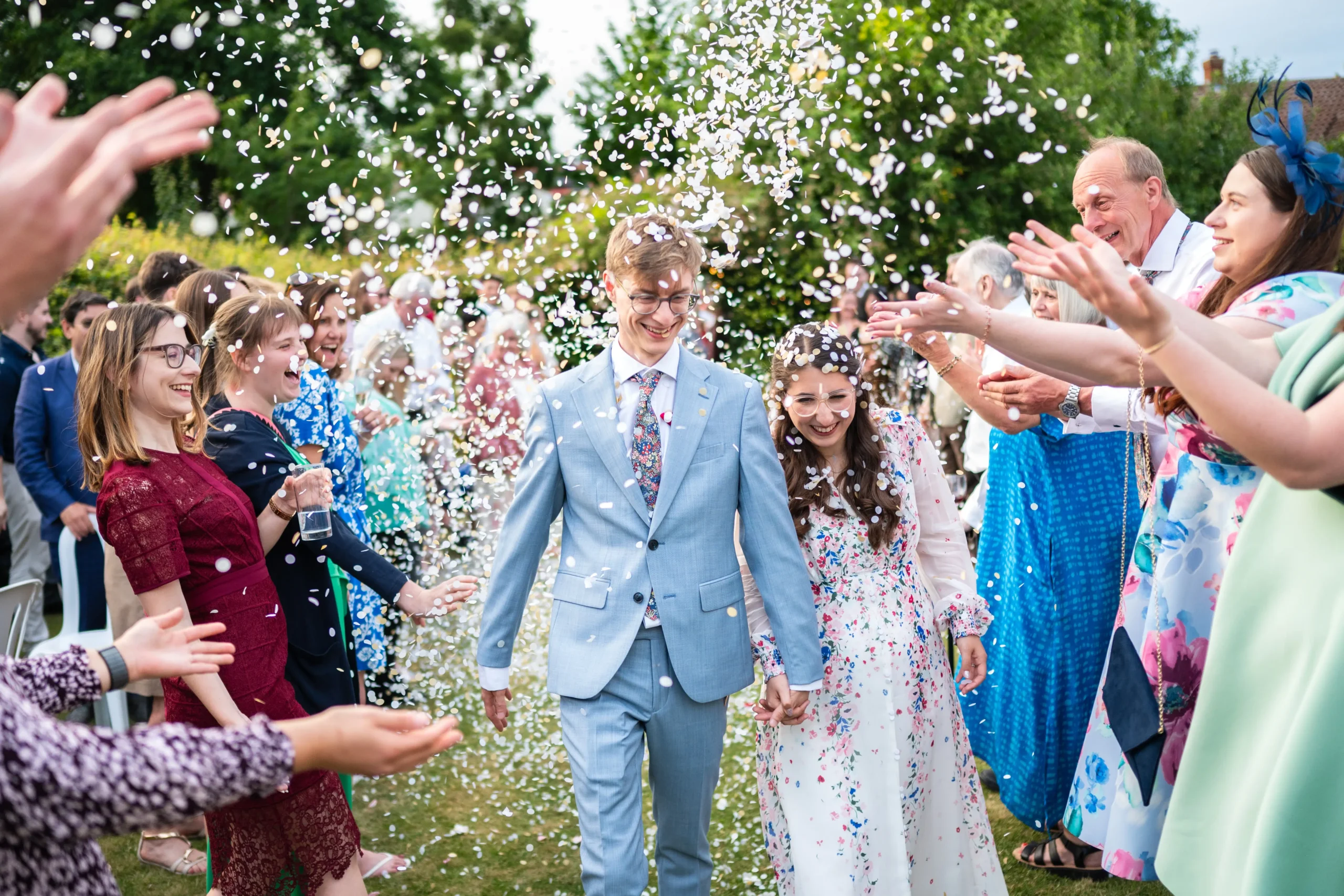 Bride in a floral wedding dress and groom in a light blue suit walking through a confetti shower with cheering wedding guests, Nottingham wedding photographer
