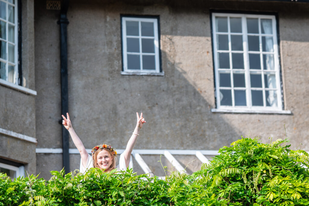Bride peeking over a garden hedge with arms raised in celebration, Nottingham wedding photographer
