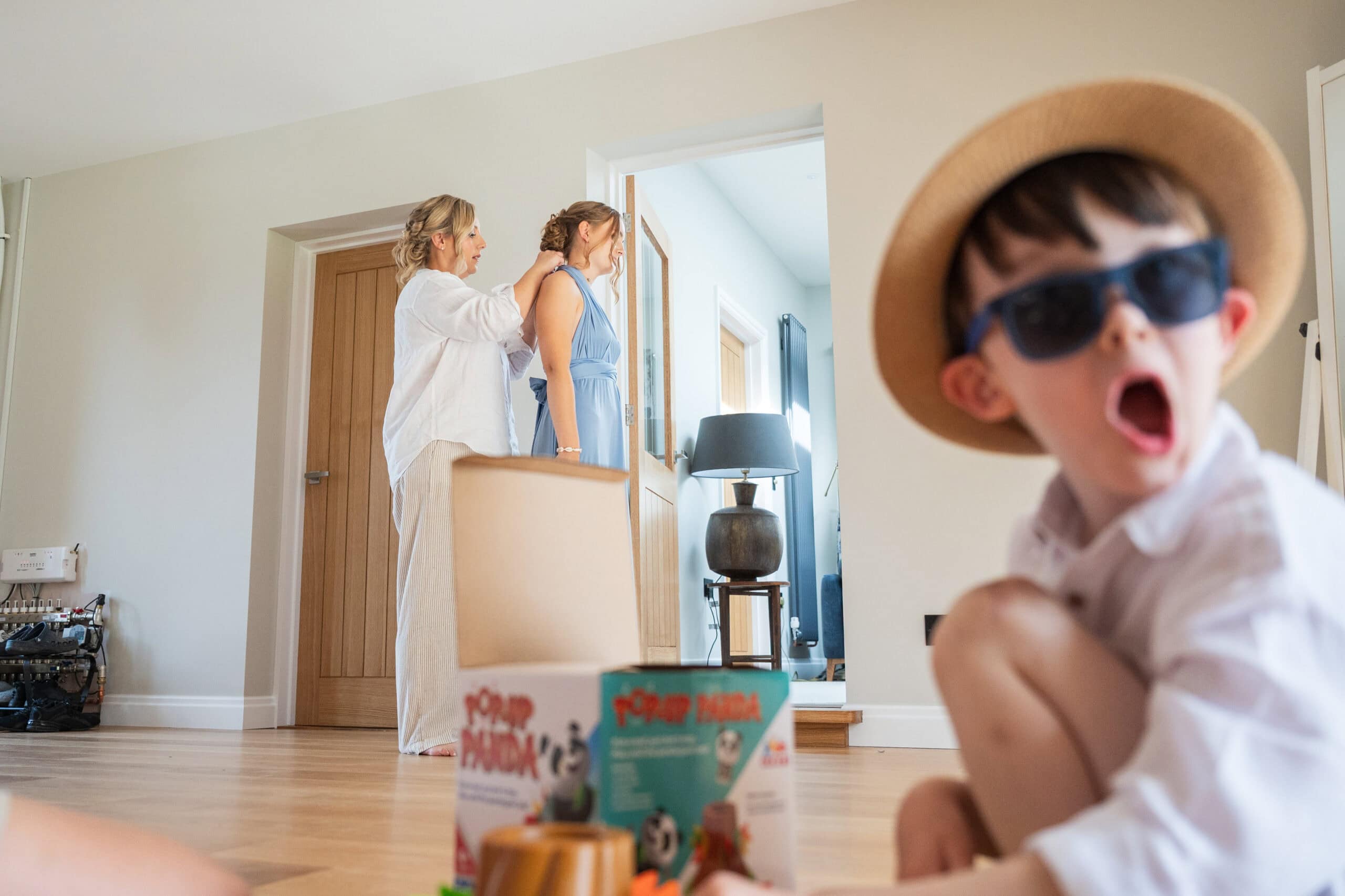 The groom's son in sunglasses and a straw hat pulls a shocked face while the bride has her hair done behind him