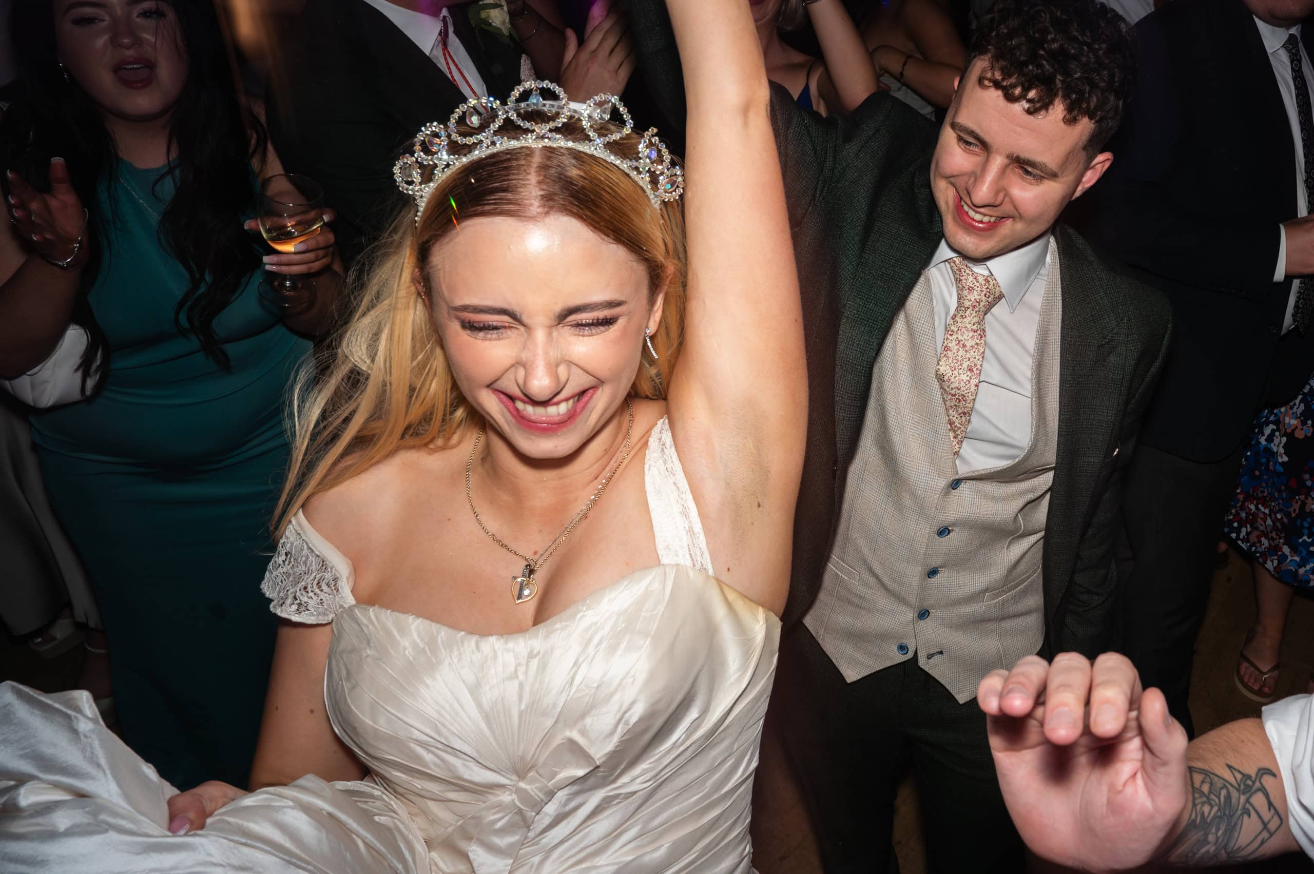 Bride in a tiara punches the air on a packed dance floor as guests celebrate around her