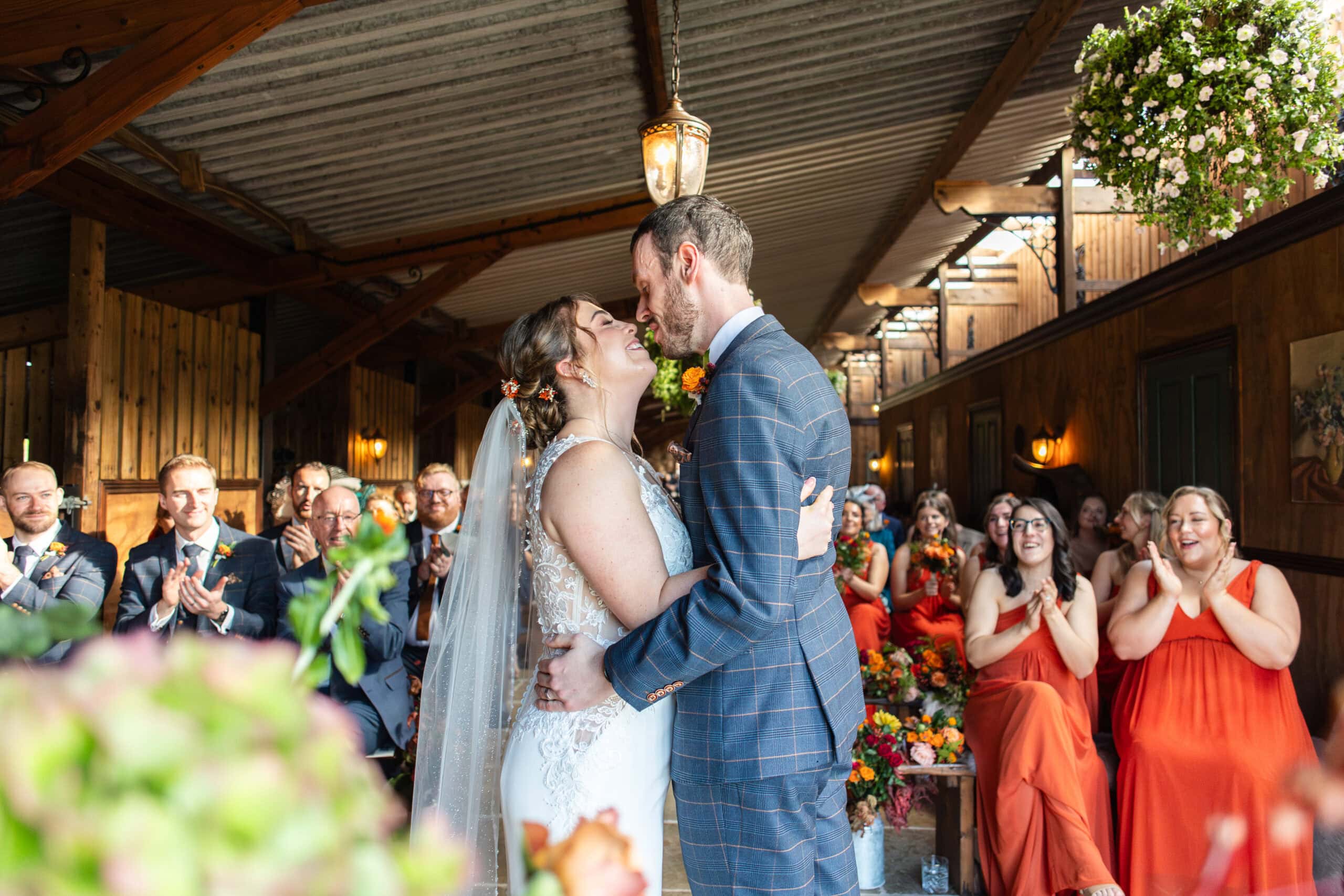 Bride and groom share their first kiss as bridesmaids in orange cheer at Willow Marsh Farm