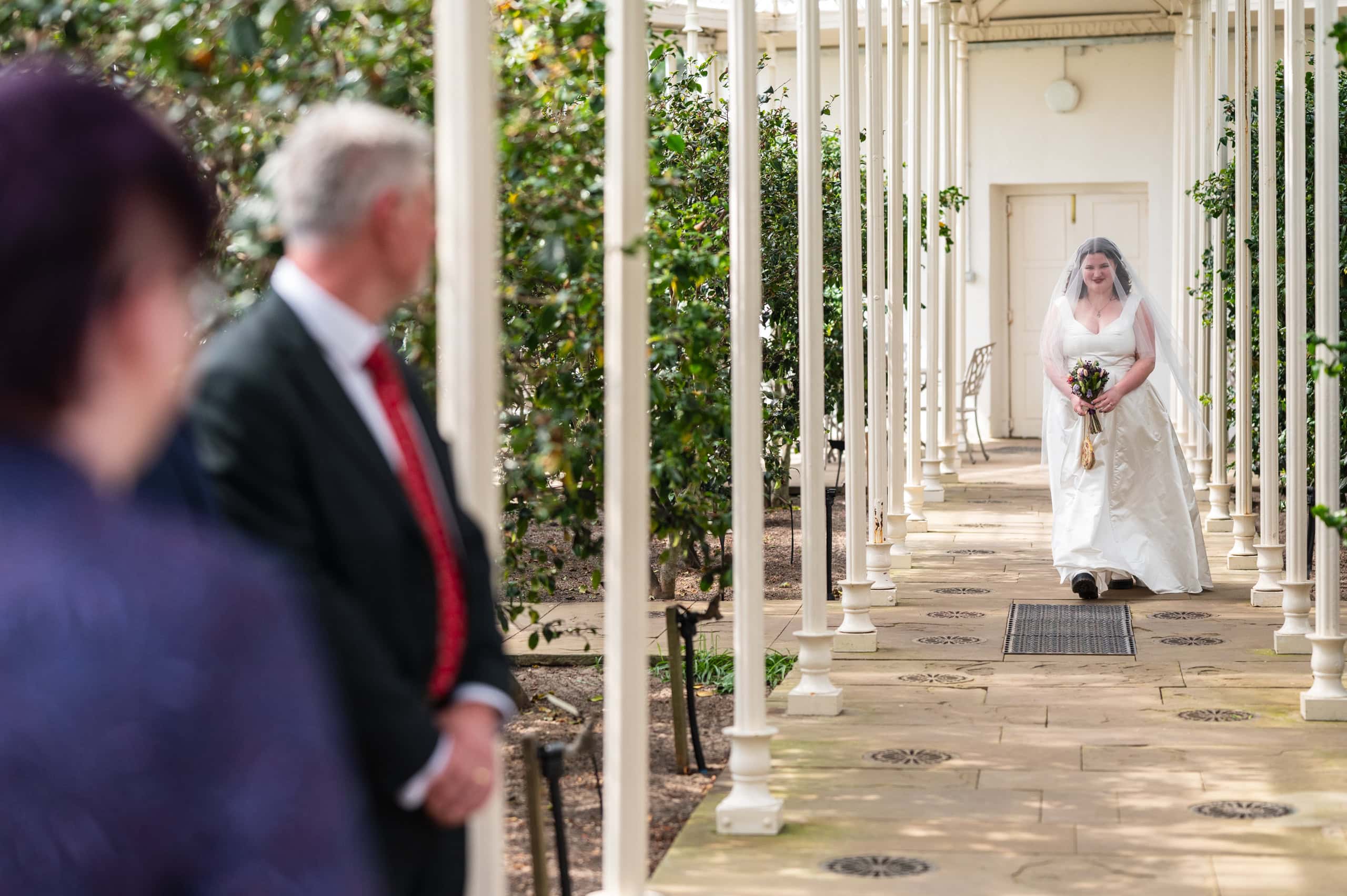 Bride walks along the colonnade of the Camellia House at Wollaton Hall towards her ceremony