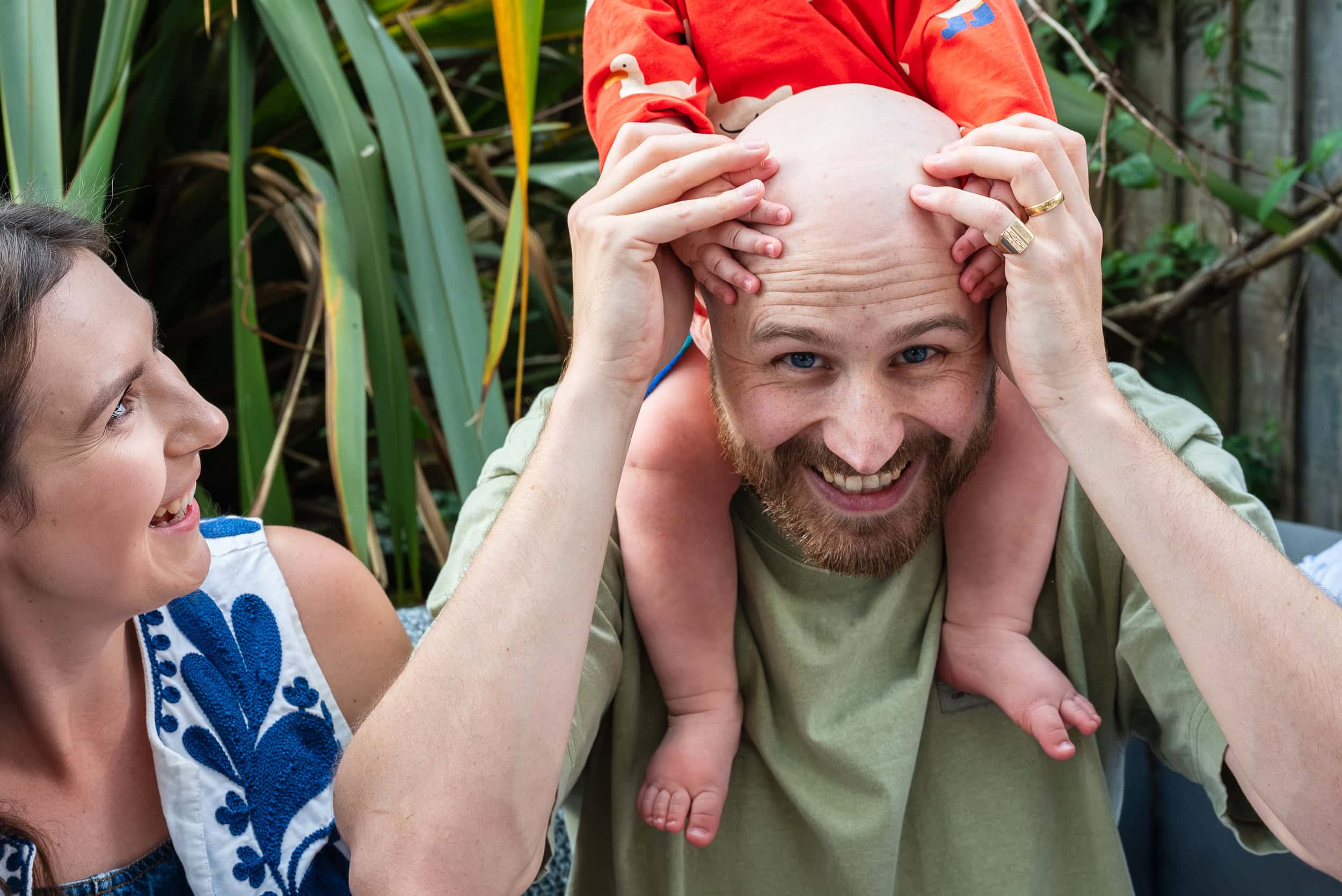 Dad laughs with his toddler on his shoulders as mum looks on