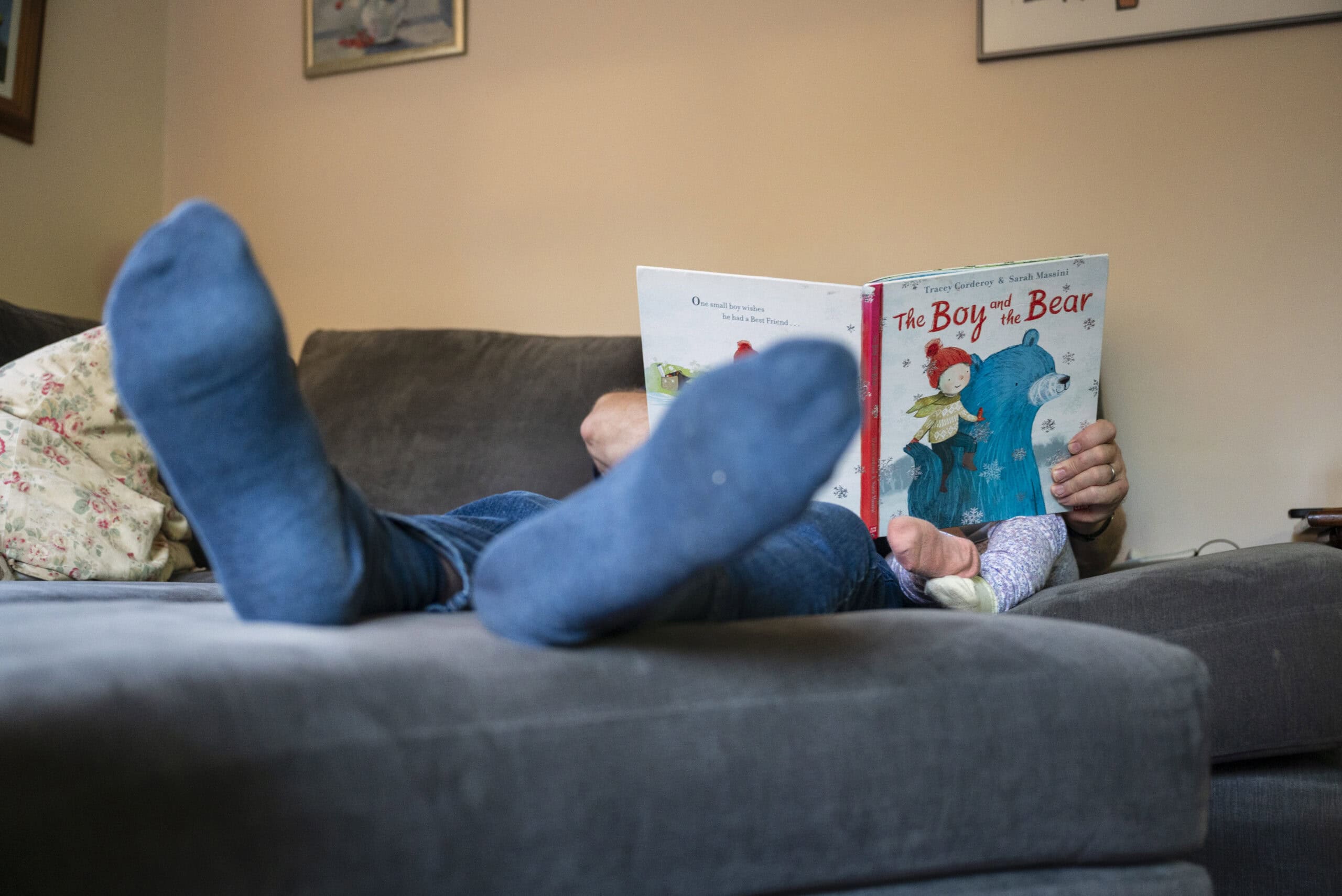 A child in blue socks lies on the sofa reading a picture book