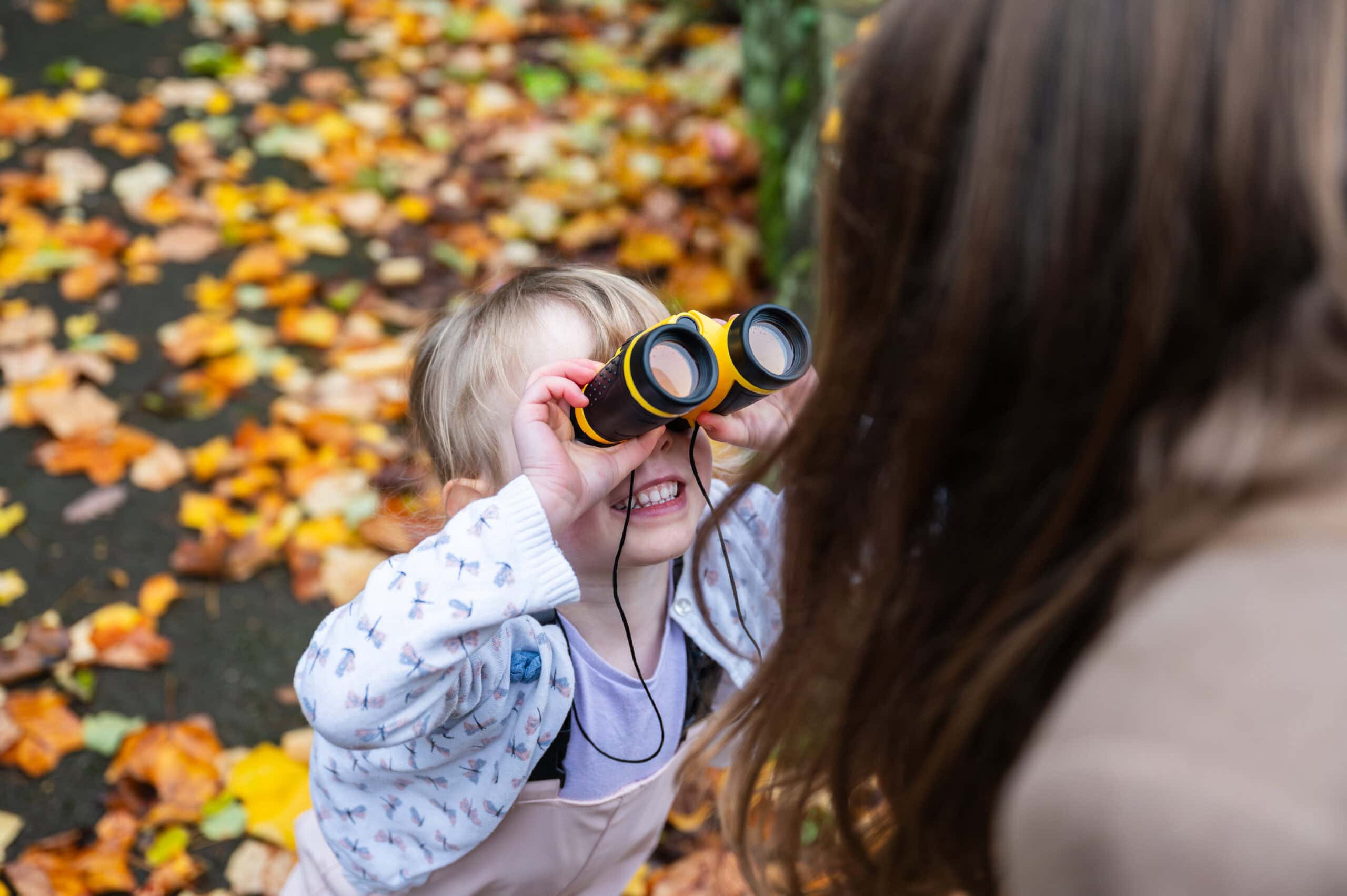 A little girl looks through toy binoculars in the autumn leaves, family photography in Nottingham