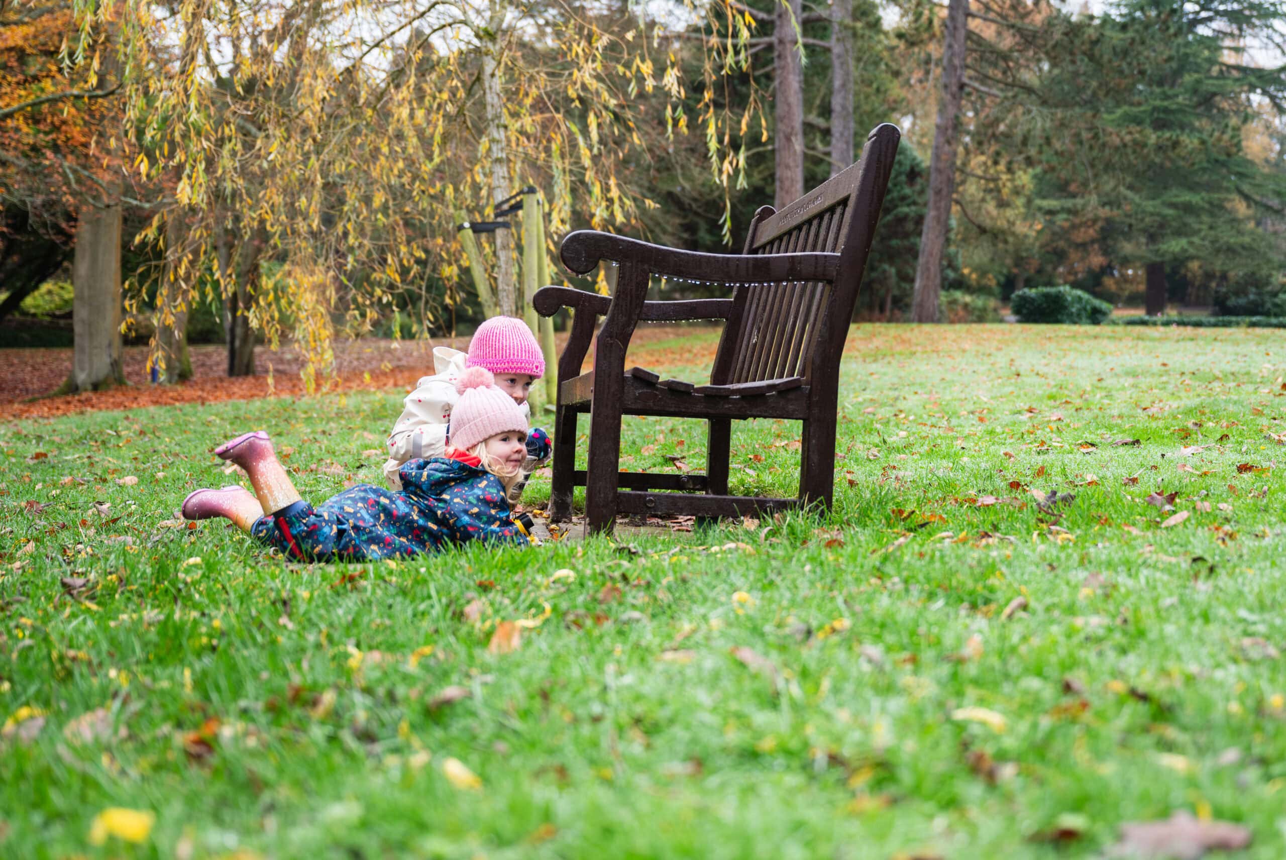 A toddler lies on the grass beside a bench in an autumn park