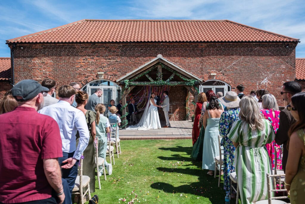 Outdoor wedding ceremony on the lawn at Dunstall Barn, guests standing either side of the aisle in the sunshine