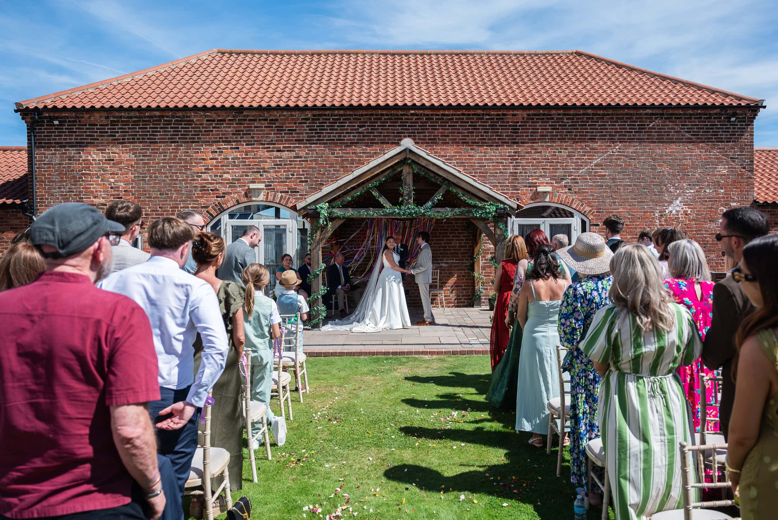 Outdoor wedding ceremony on the lawn at Dunstall Barn, guests standing either side of the aisle in the sunshine