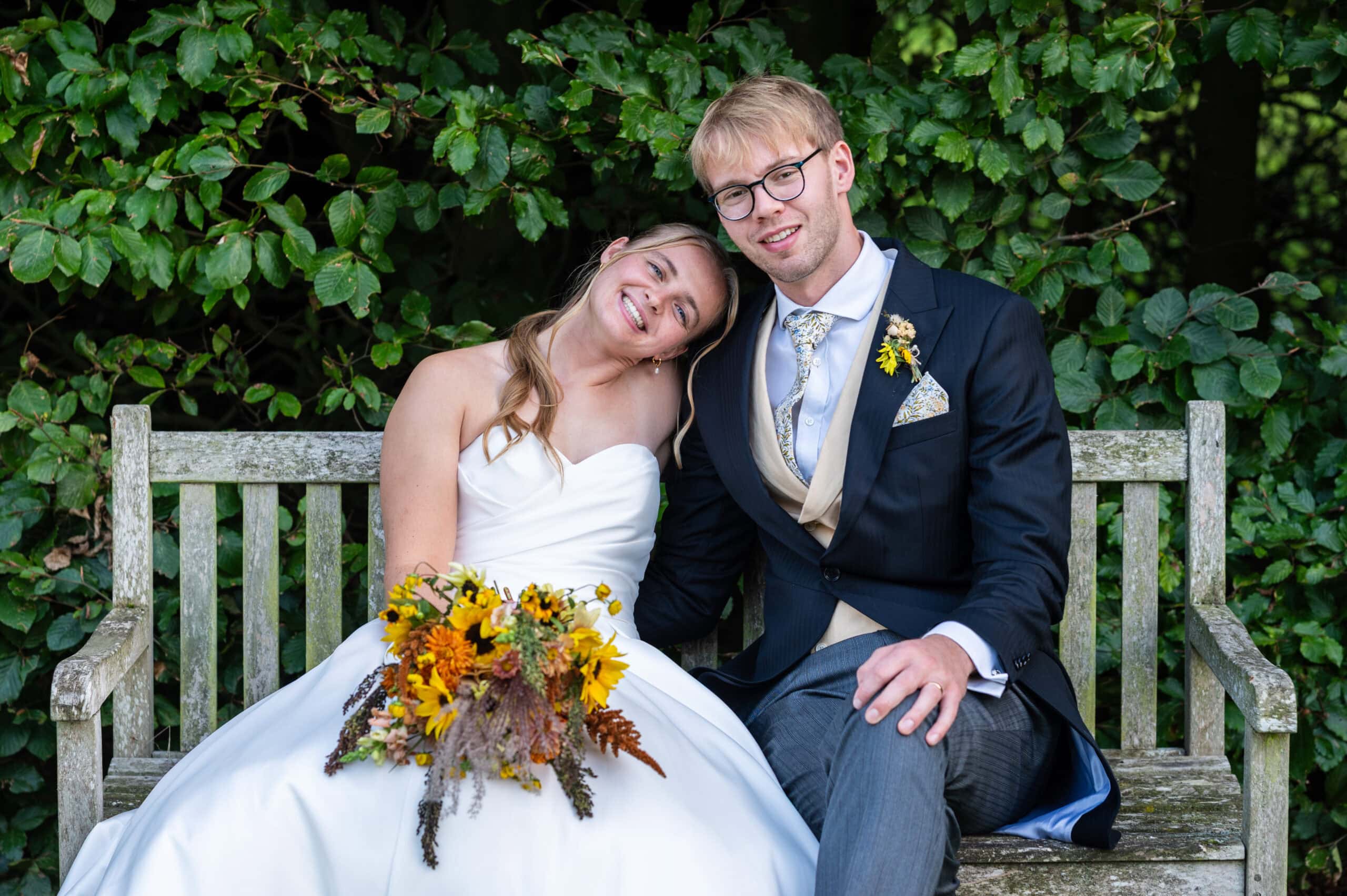 A bride and groom laughing on a wooden bench against the hedges at Holme Pierrepont Hall, sunflower bouquet in her lap.