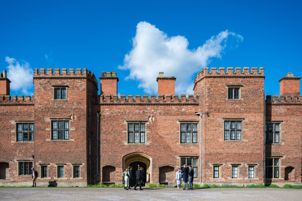 The red-brick front of Holme Pierrepont Hall under a blue sky, wedding guests gathered by the entrance.