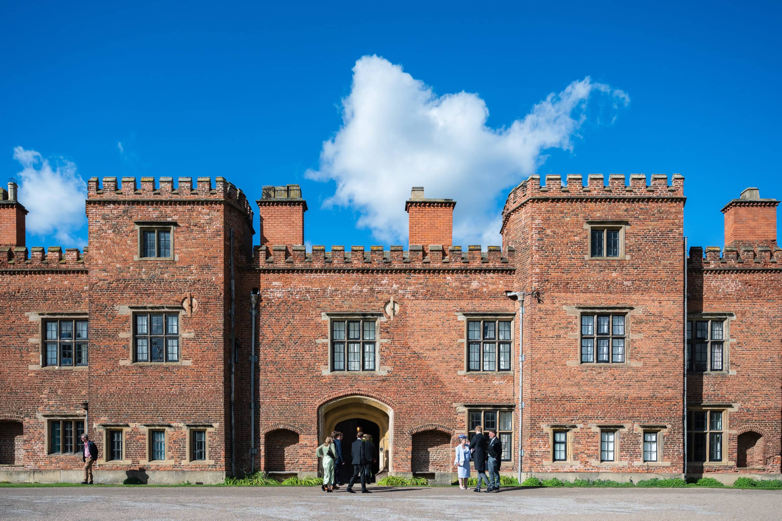 The red-brick front of Holme Pierrepont Hall under a blue sky, wedding guests gathered by the entrance.