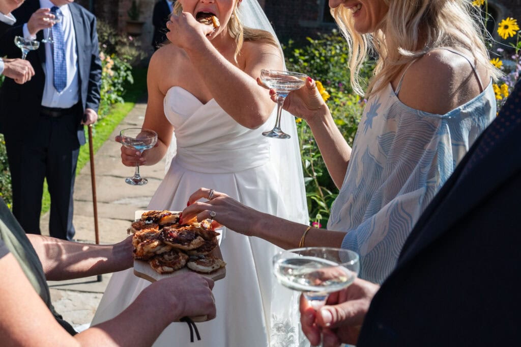 A bride laughing with friends in the courtyard garden at Holme Pierrepont Hall as a plate of canapés is passed round.