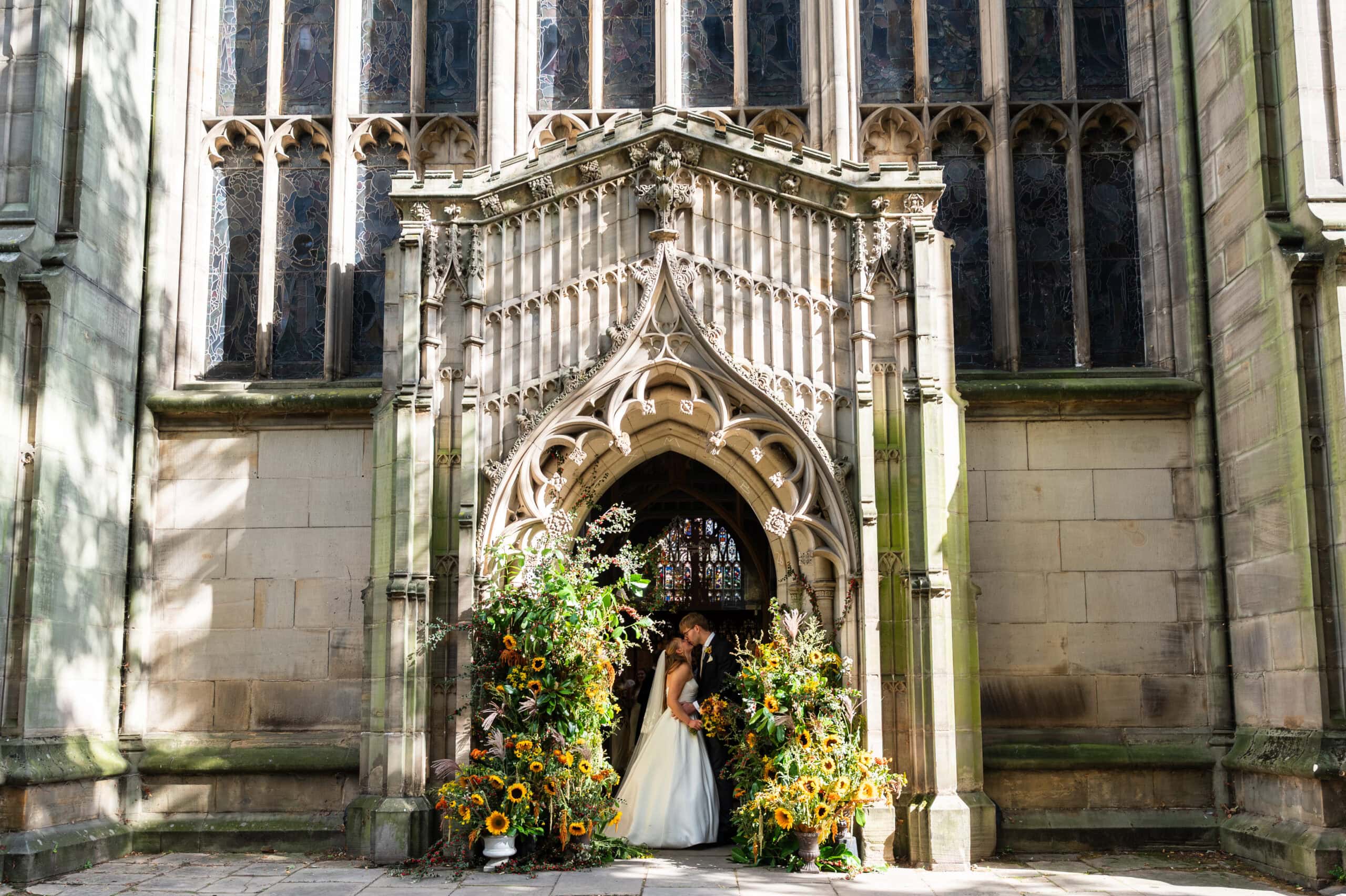 A bride and groom kissing in the carved stone doorway of St Mary's in the Lace Market, framed by sunflower arrangements.