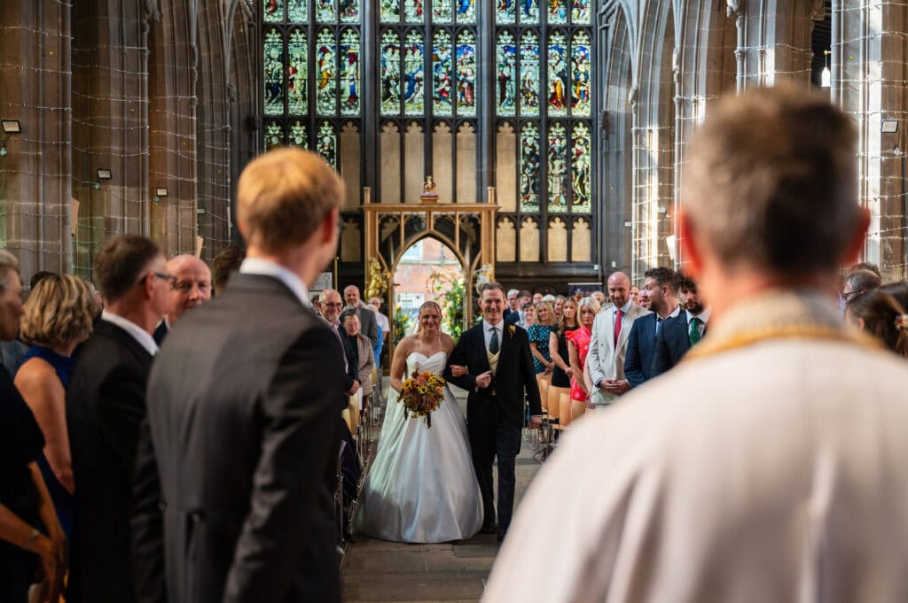 A bride walking down the aisle of St Mary's in the Lace Market towards the groom, the stained glass behind them.