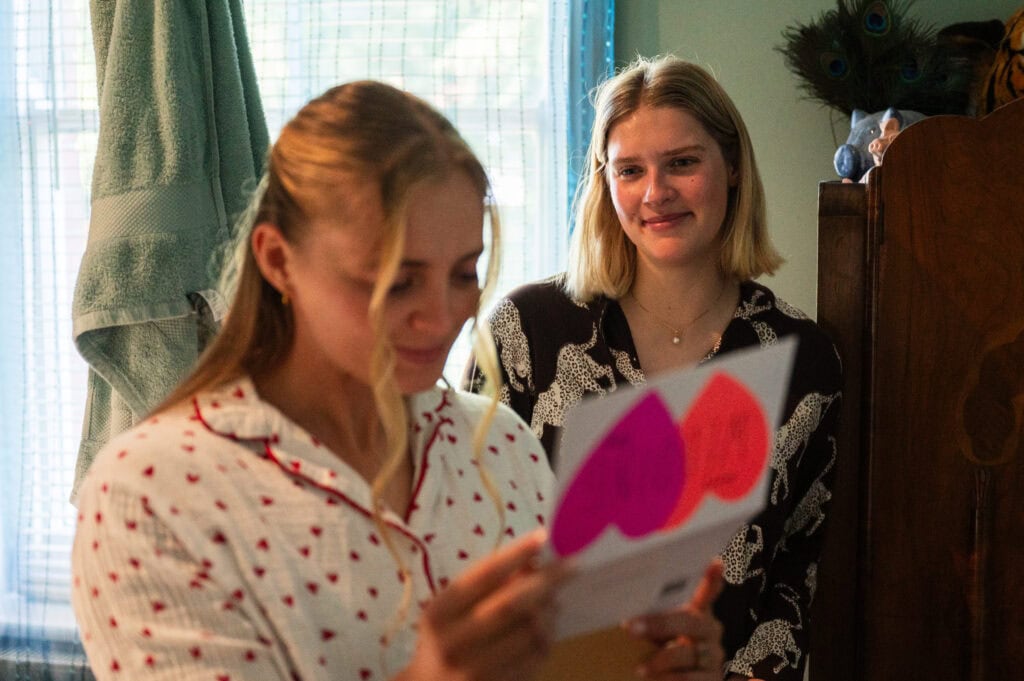 A bride in spotted pyjamas reading a heart card from her bridesmaid, who watches beside her.