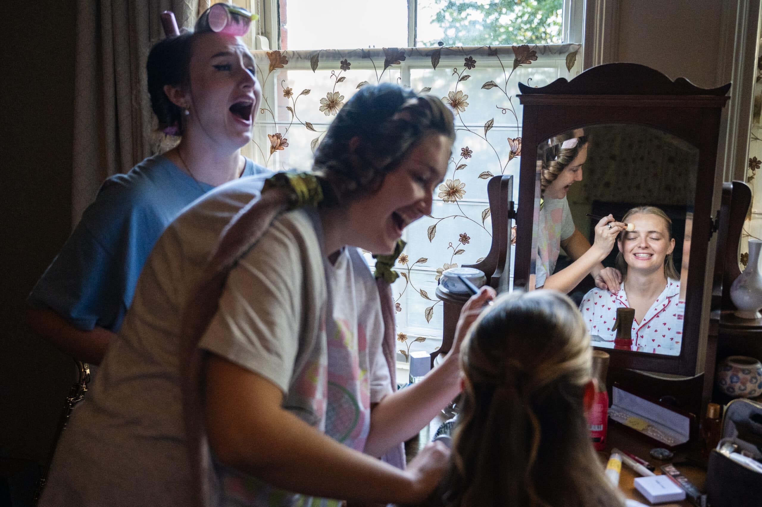 Bridesmaids laughing by a dressing-table mirror while the bride's hair is done on the wedding morning.