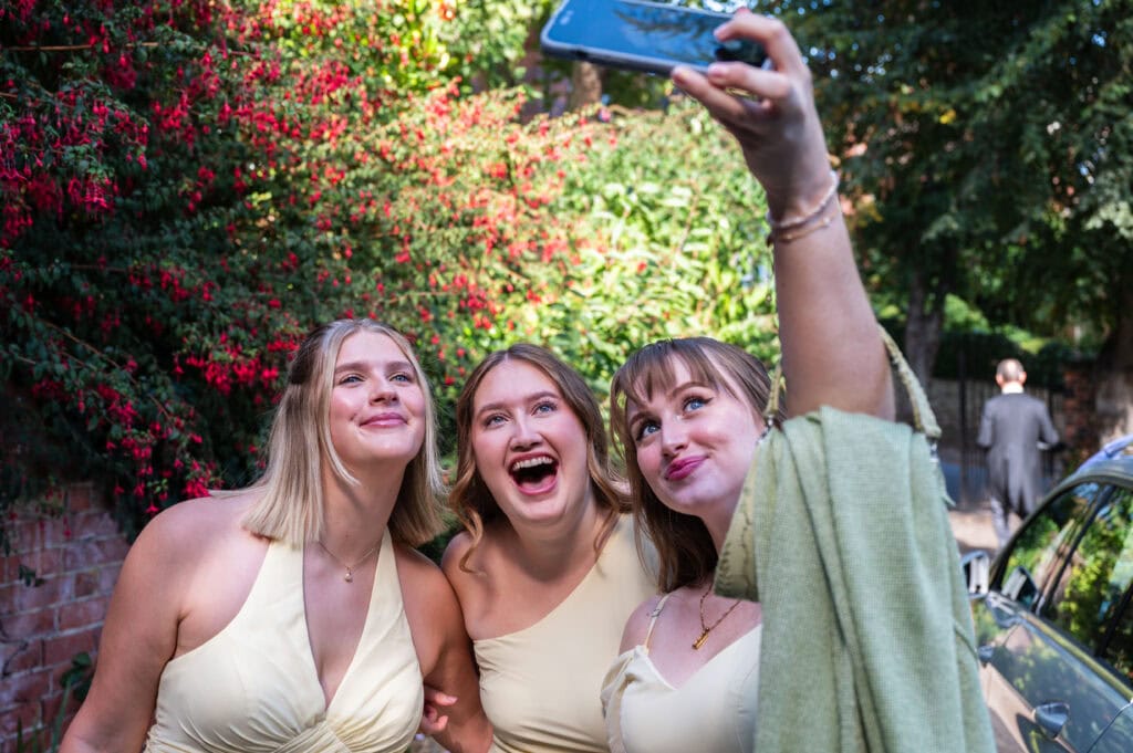 Three bridesmaids in pale yellow dresses pulling faces for a selfie outside the church.