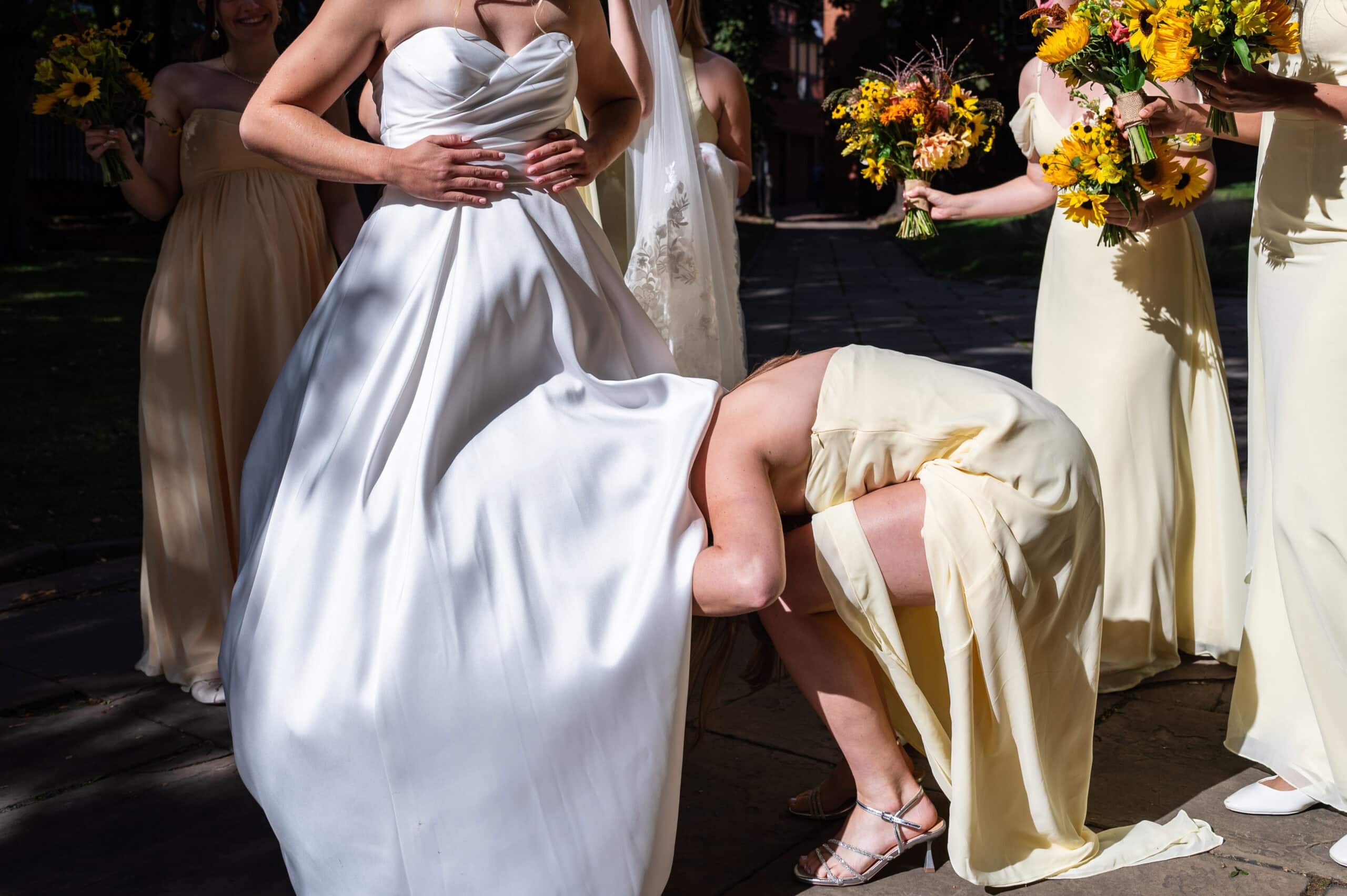 A bridesmaid in a pale yellow dress bent right under the bride's skirt outside St Mary's church, sunflower bouquets around them.