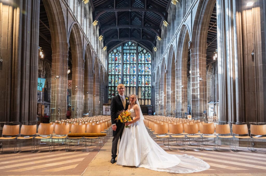 A bride and groom standing in the empty nave of St Mary's in the Lace Market under a large stained glass window.