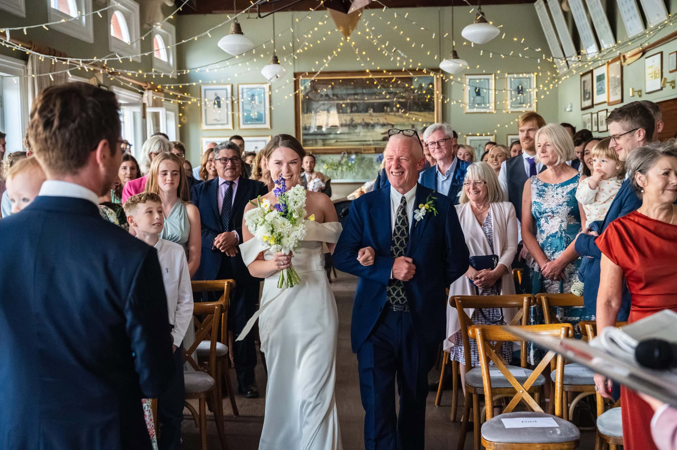 Ella walking down the aisle with her father during her wedding ceremony in the Long Room at London Rowing Club, fairy lights strung across the ceiling