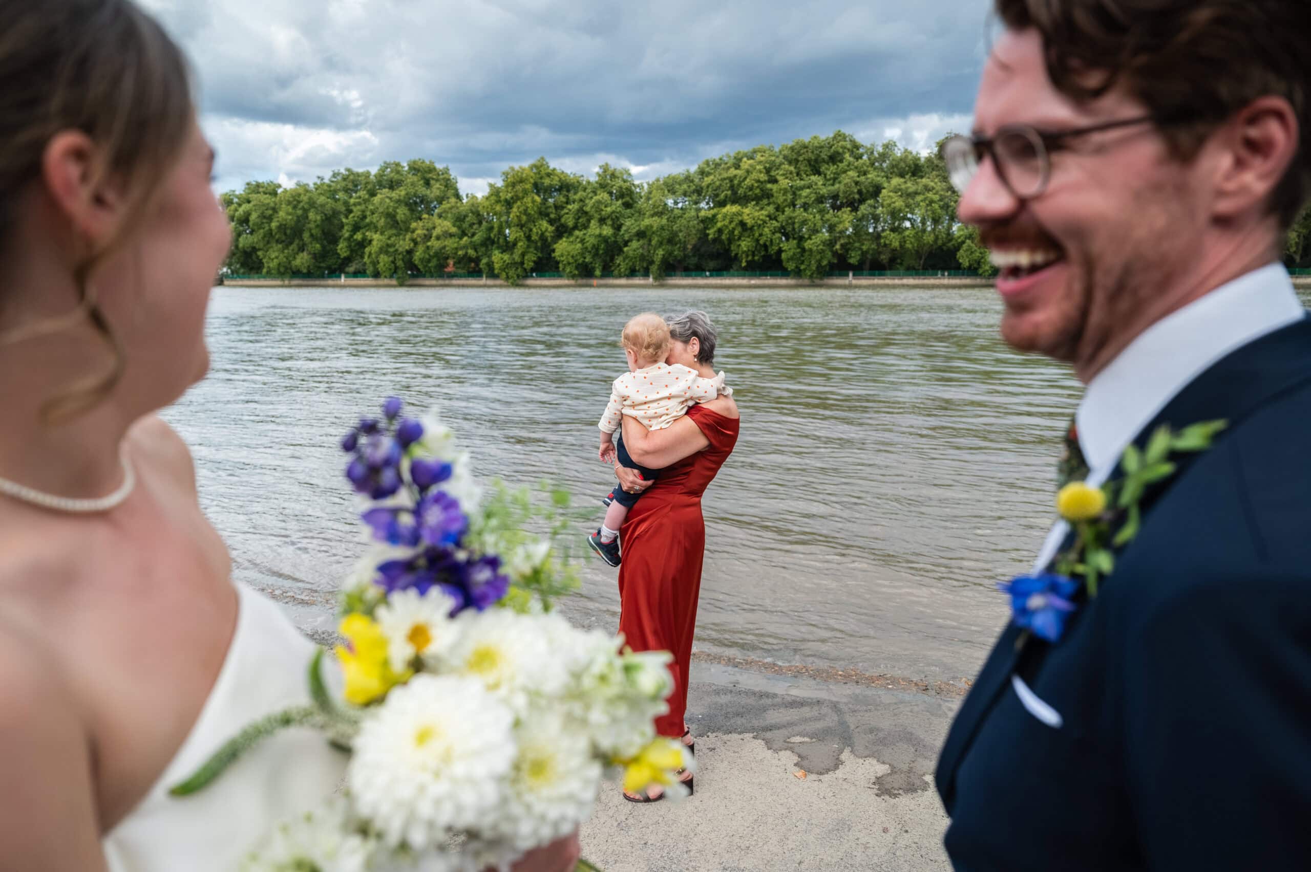 A wedding guest in a red dress holding a small child beside the River Thames outside London Rowing Club