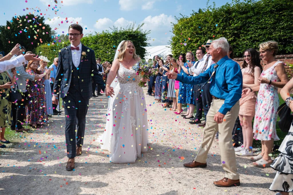 Confetti thrown over a bride and groom walking the wide path out of Pippley Walled Garden, guests lining both sides
