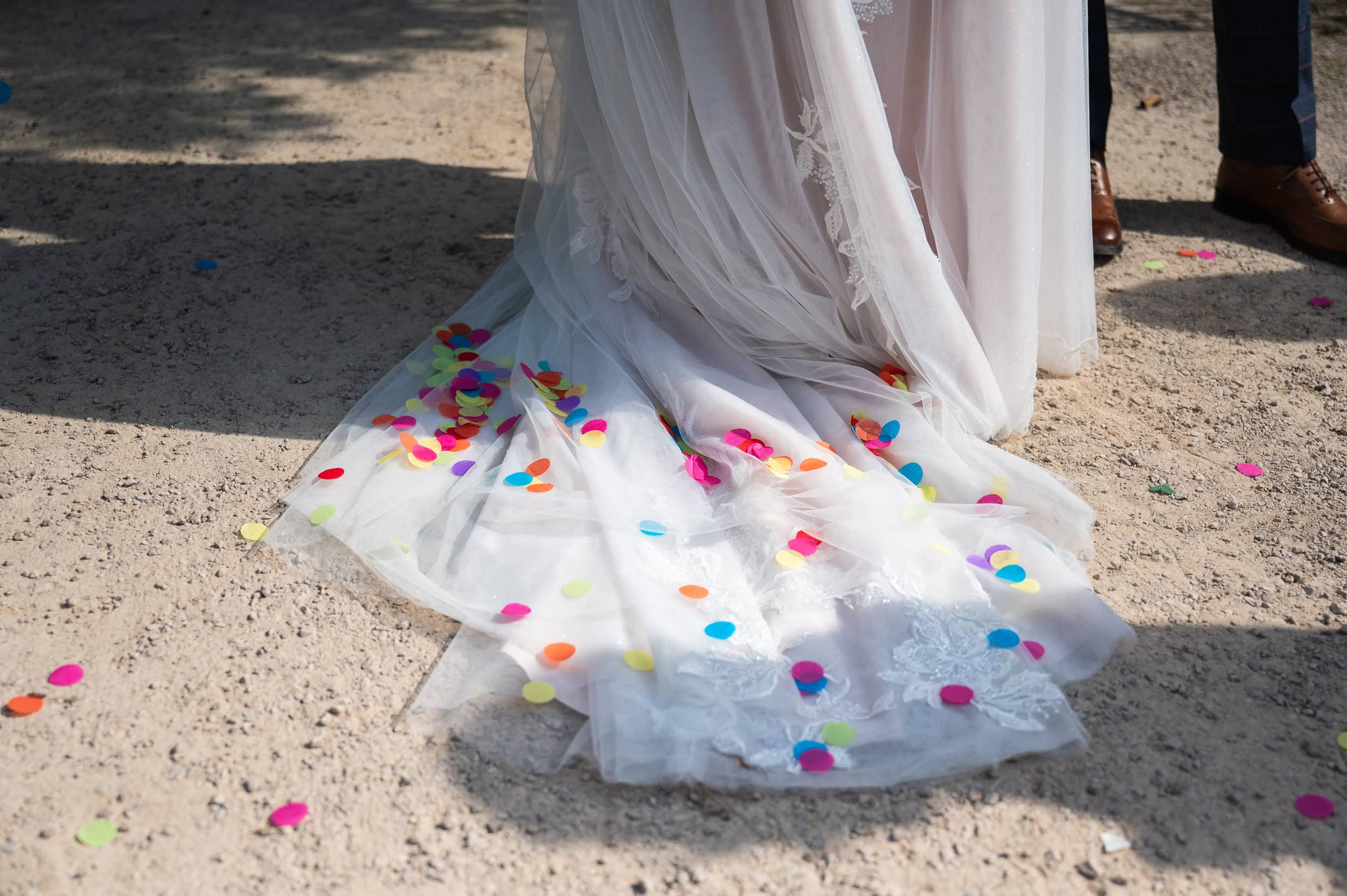 Bright confetti scattered across a bride's tulle train on the gravel path