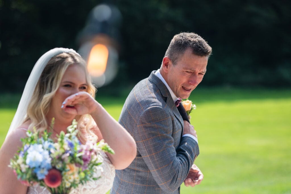 Groom doubled over laughing beside his bride, who is wiping her eye, on the lawn