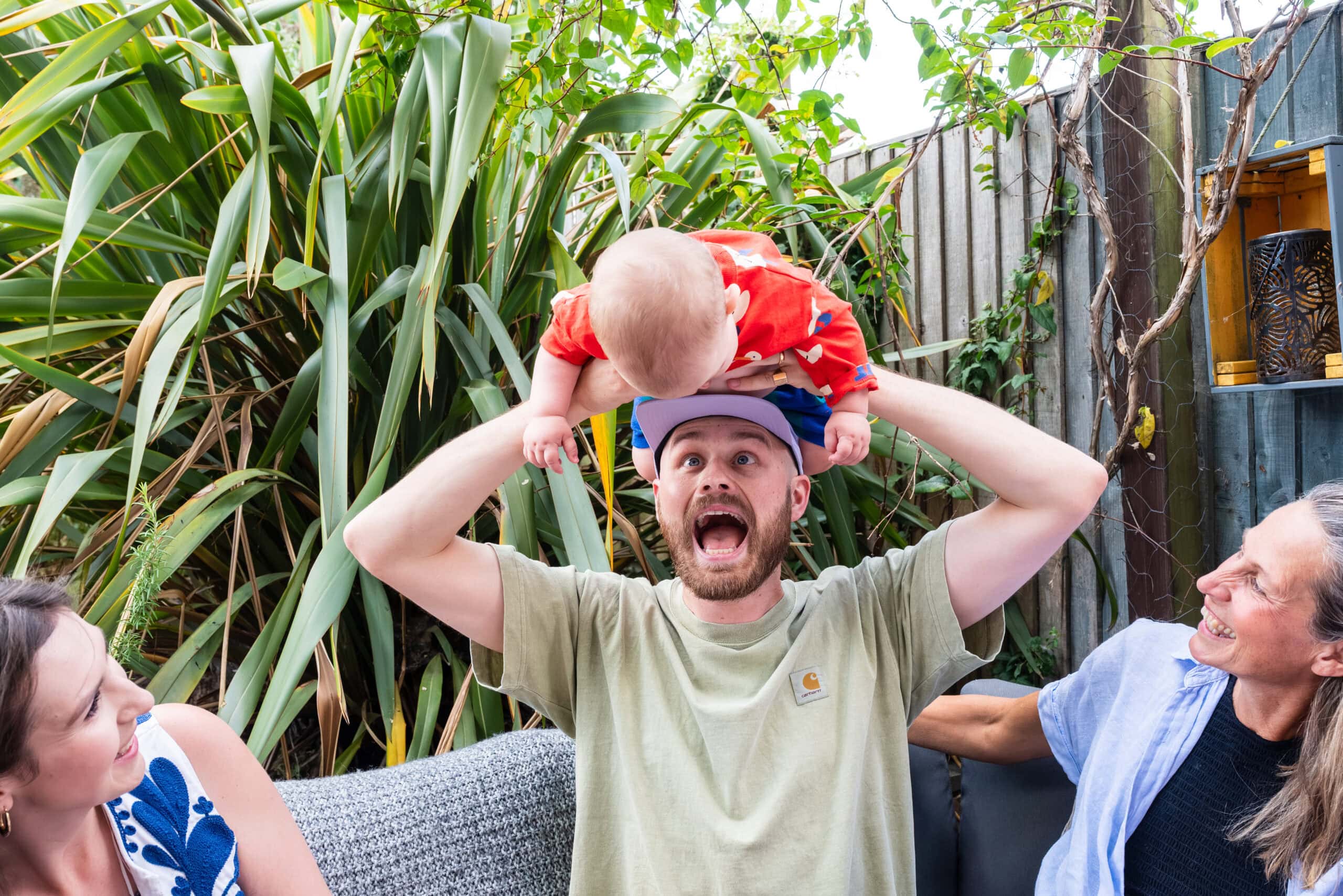 Dad lifts his baby onto his head in the back garden while the family laughs, at-home family photography