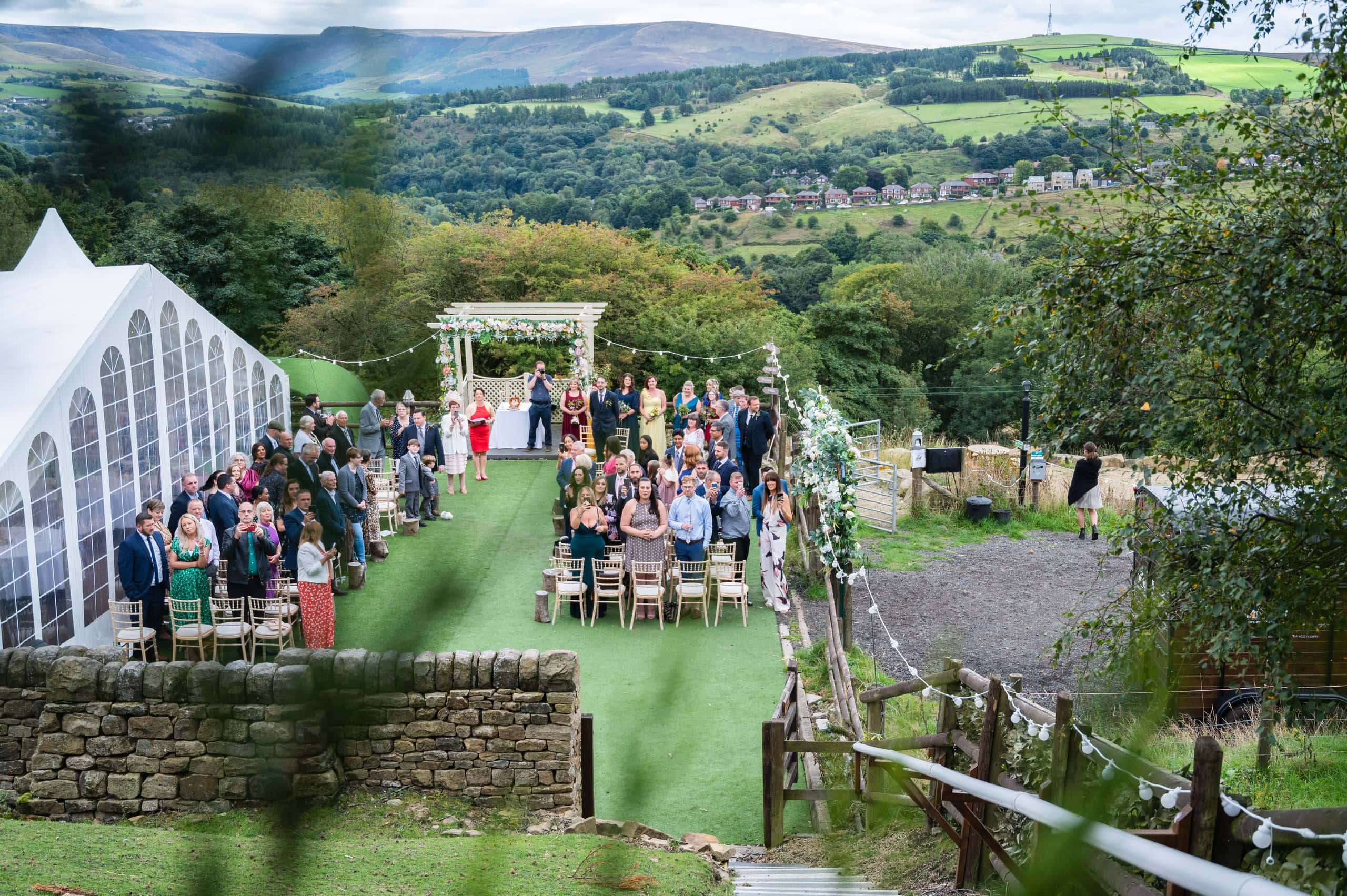 Outdoor celebrant wedding ceremony on a hillside lawn with views over the valley at The Stables Wedding Farm, Oldham